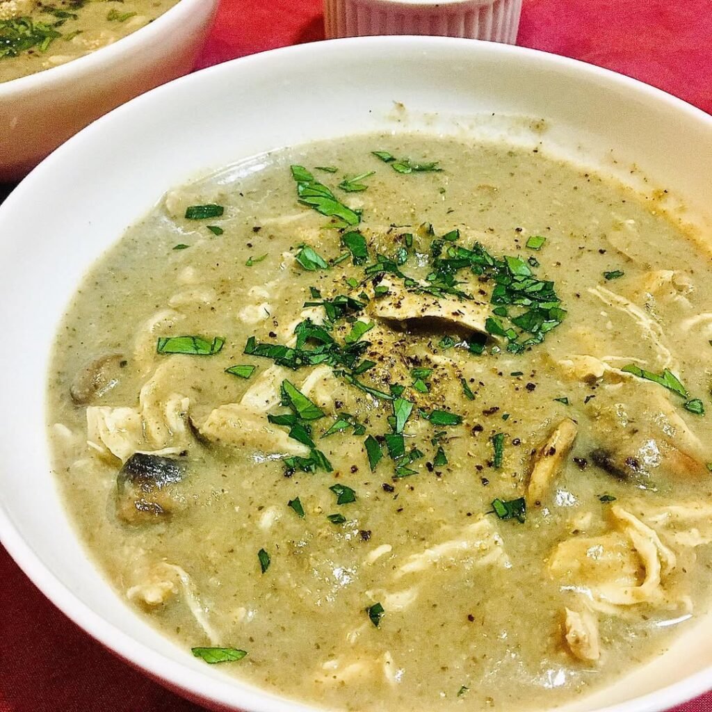 A bowl of chicken and mushroom soup with chunks of chicken, sliced mushrooms, and a slice of bread on the side.