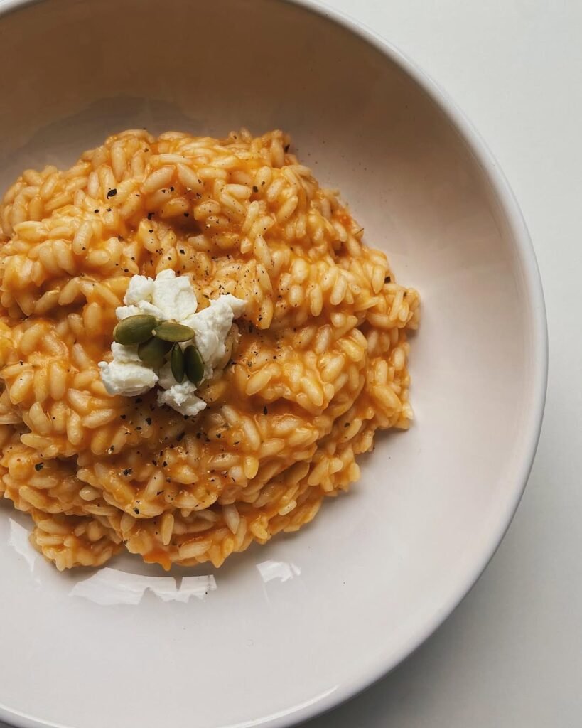 7 Fall Dinner Ideas To Warm Up Your Evenings 5 A bowl of creamy pumpkin risotto topped with fresh herbs and Parmesan cheese, with a small pumpkin in the background.