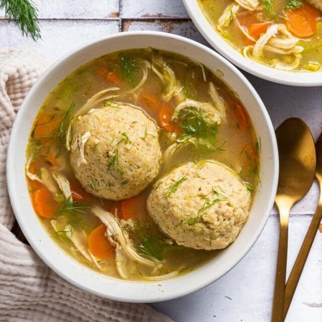 A bowl of classic Jewish chicken soup with matzo balls, garnished with dill, served with bread on the side.