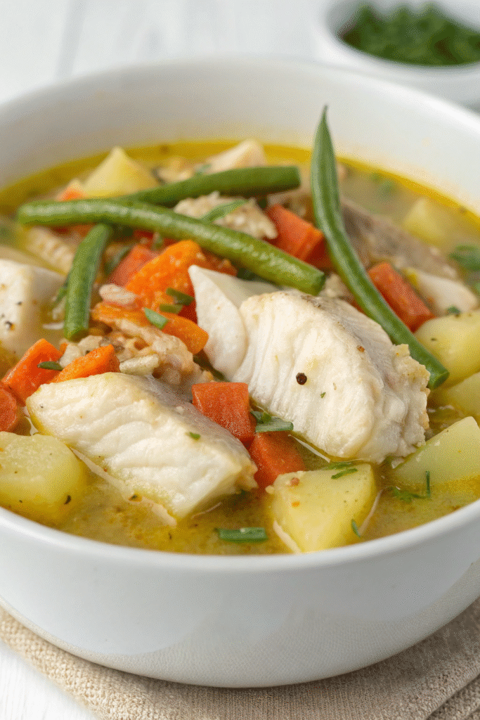 A bowl of fish and vegetable chowder with a spoon and fresh herbs